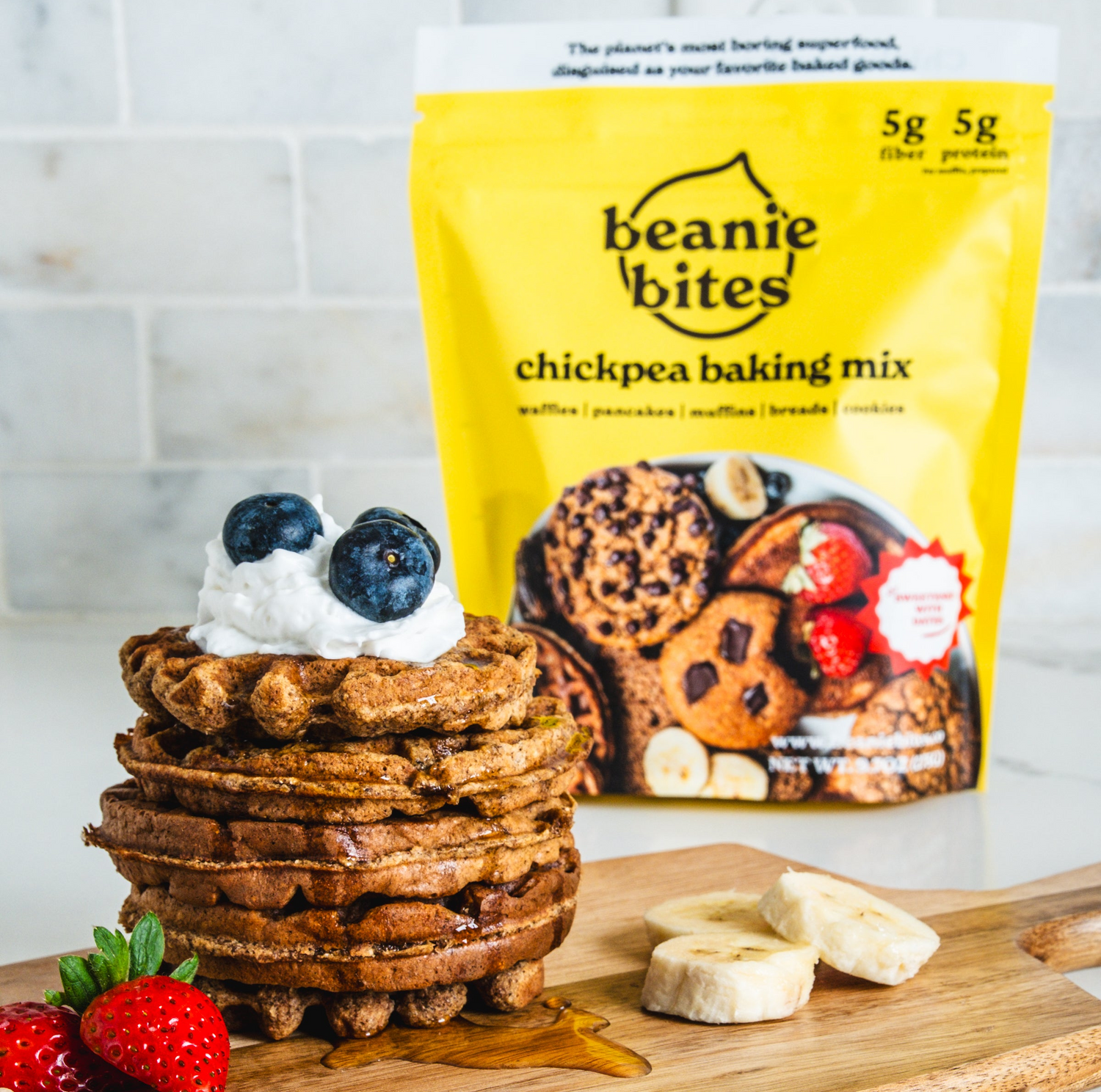 Stack of pancakes with blueberries and whipped cream on a wooden board, with a bag of beanie bites chickpea baking mix in the background.