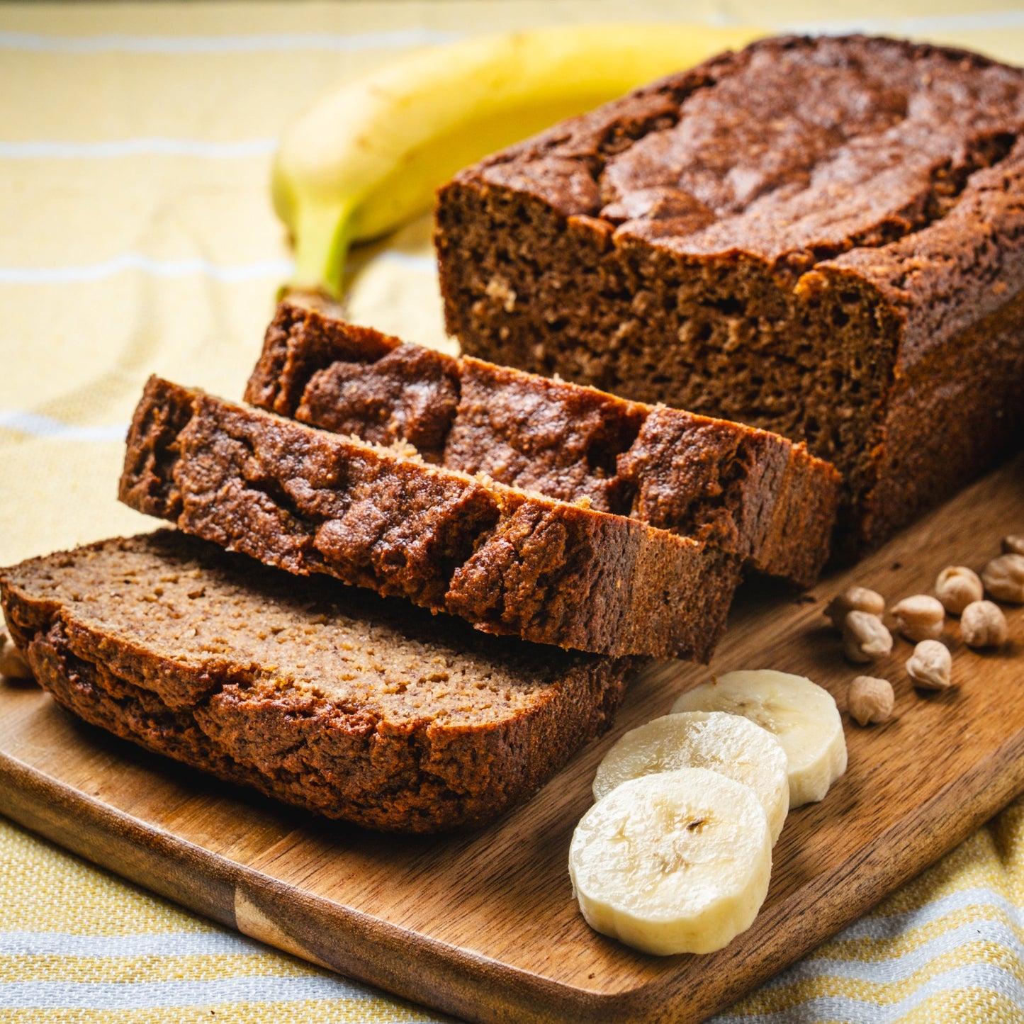 Loaf of banana bread with slices on a wooden cutting board, surrounded by bananas and chickpeas on a striped fabric background. made with chickpeas and sweetened with dates 
